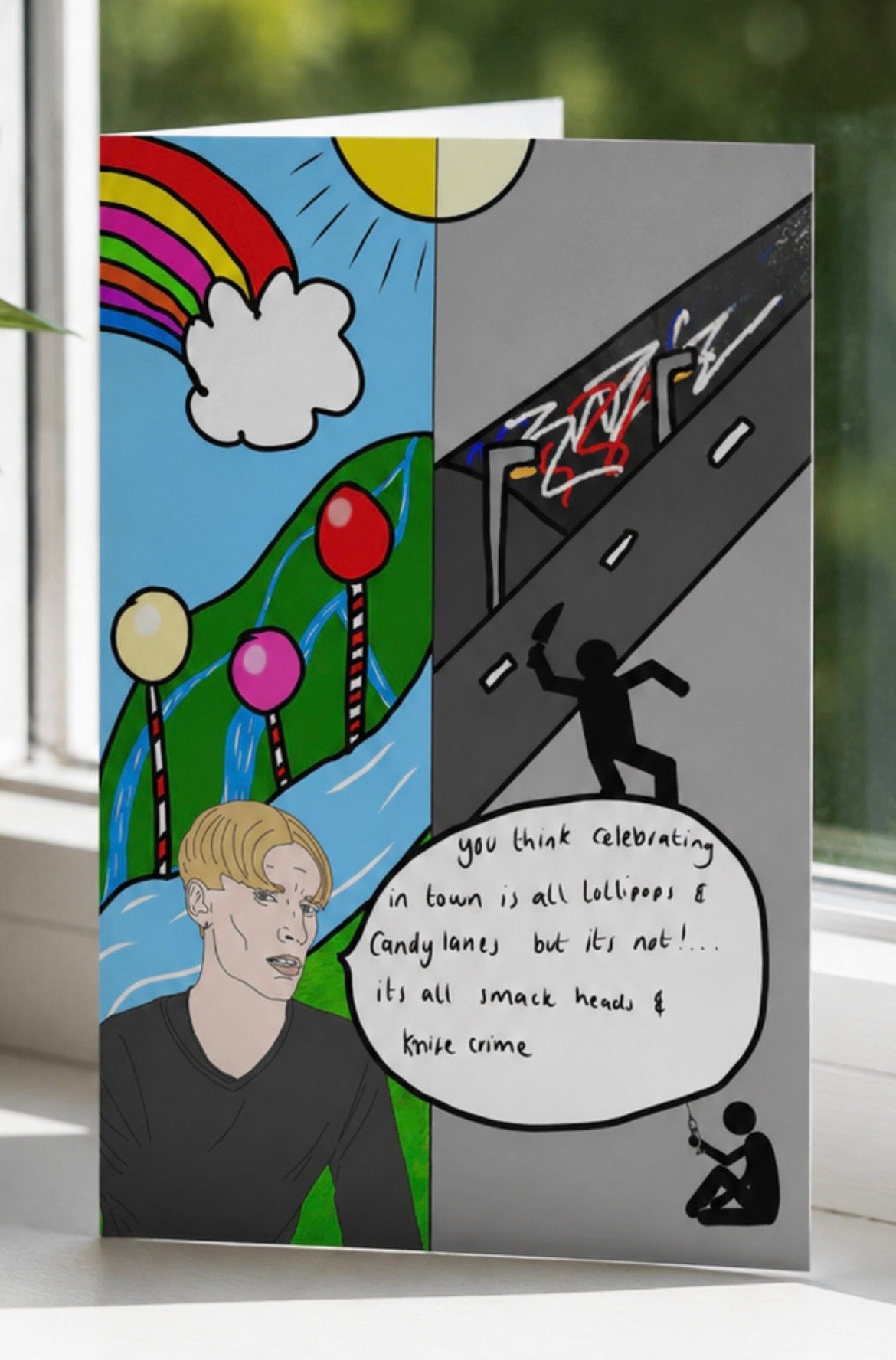 A split-panel greeting card standing on a windowsill, showing a colorful cartoon landscape on the left and a dark street-art scene on the right, with a speech bubble containing handwritten text.
